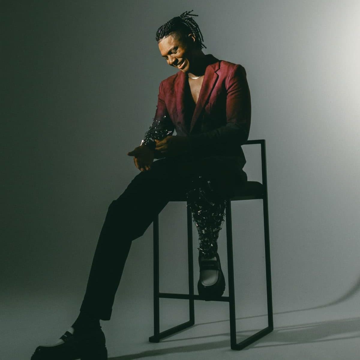 A maroon suede blazer over black tailoring with beaded detailing, seated on a high stool under a single light
