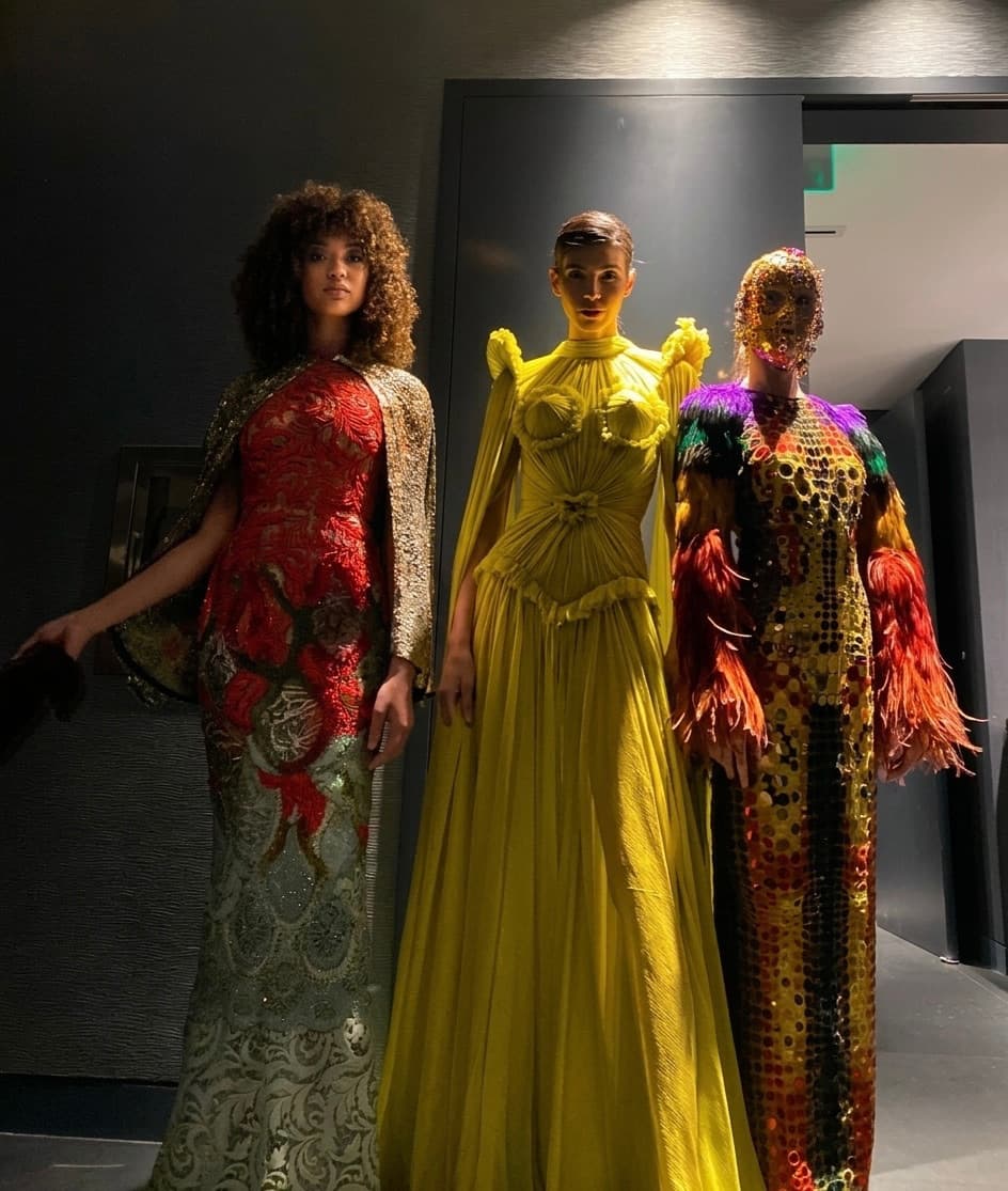Three models backstage in a red and gold brocade gown, a chartreuse draped gown, and a sequinned feather look