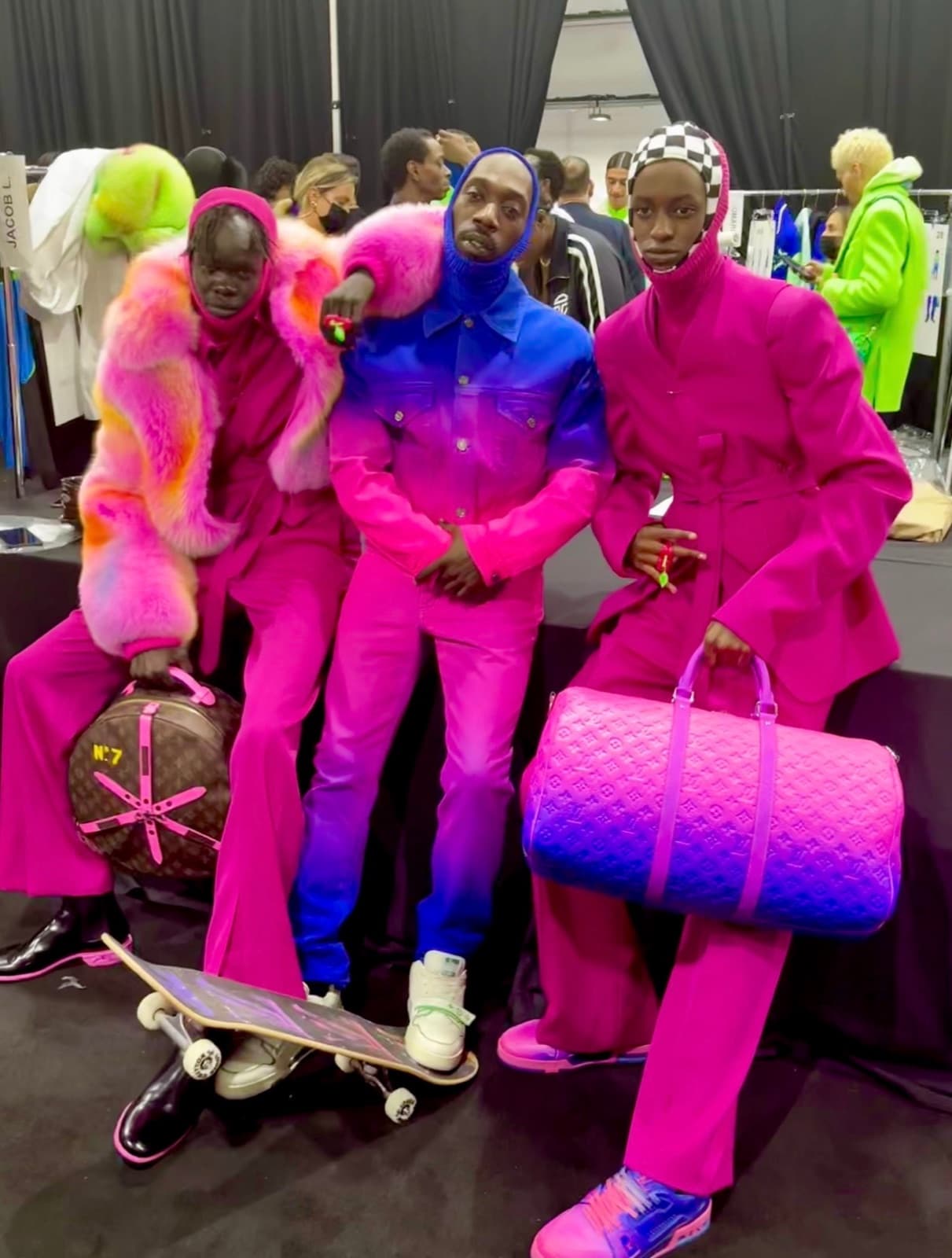 Three models backstage in fuchsia and cobalt tailoring, holding monogram duffle bags