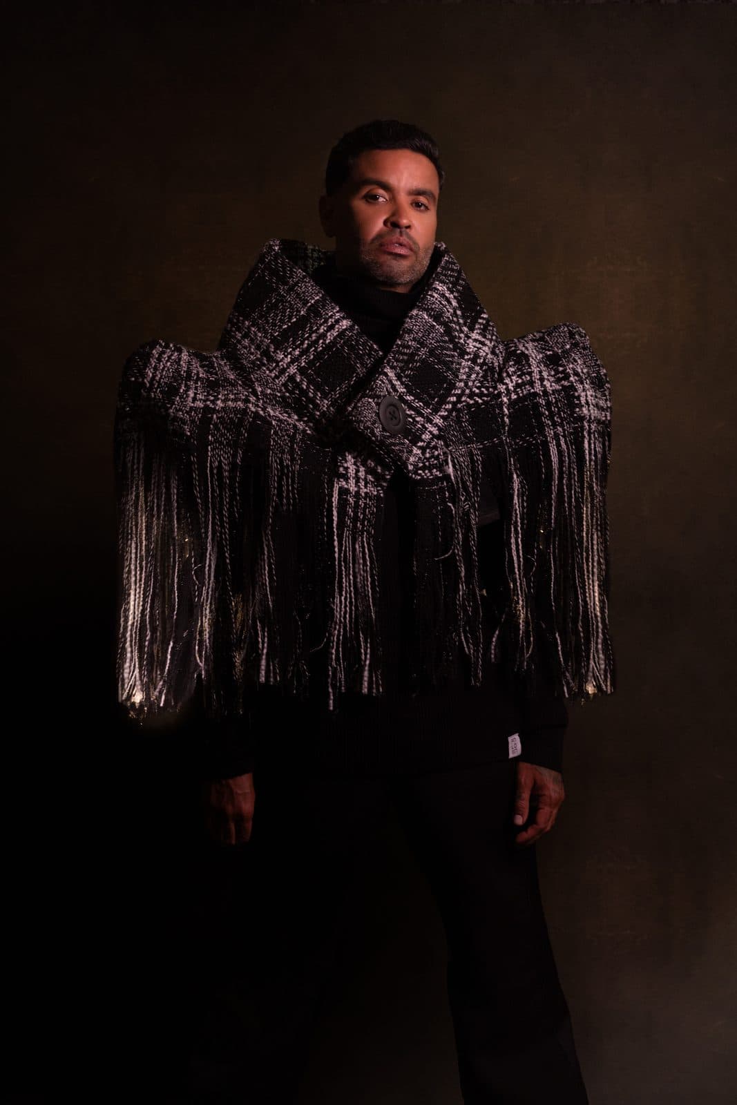 A fringed black and white tweed shawl worn over a black knit, lit low against a dark ground