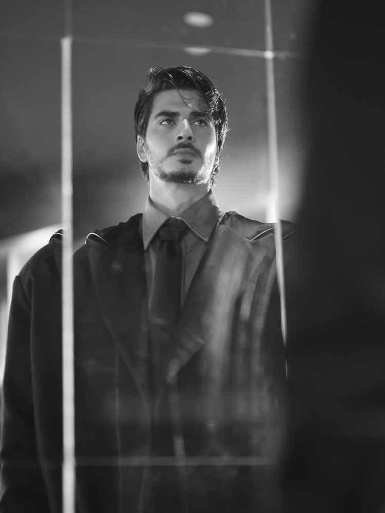 Black and white editorial portrait — a man in a dark tailored coat and tie reflected in glass