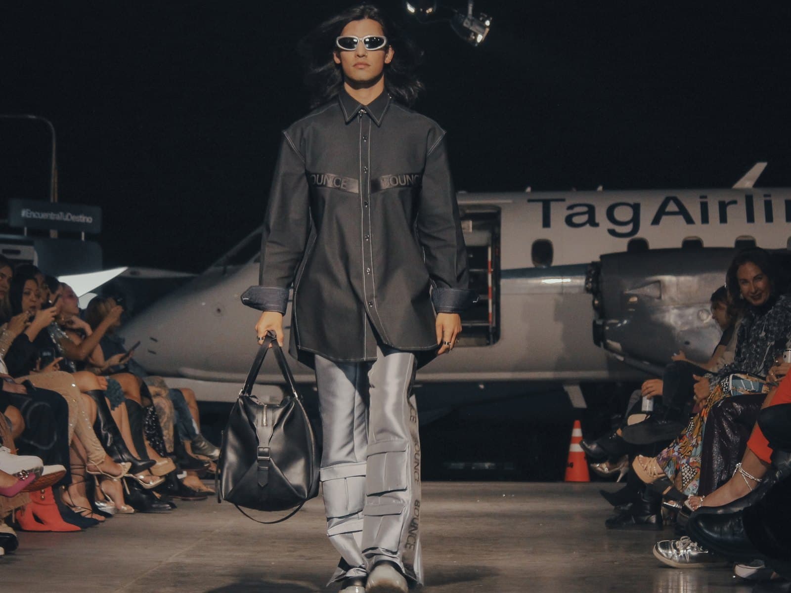 Runway look — a topstitched charcoal overshirt with silver cargo trousers, walked past a parked aircraft