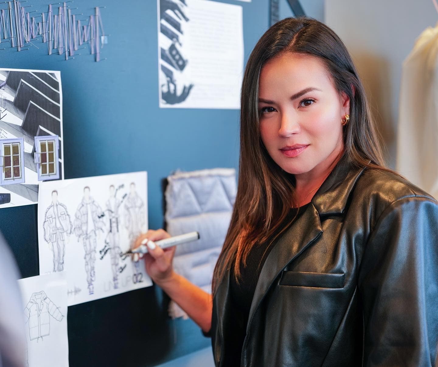 Paloma Balvin at the studio wall in a black leather jacket, marker in hand, annotating a sketched menswear line-up pinned beside fabric swatches