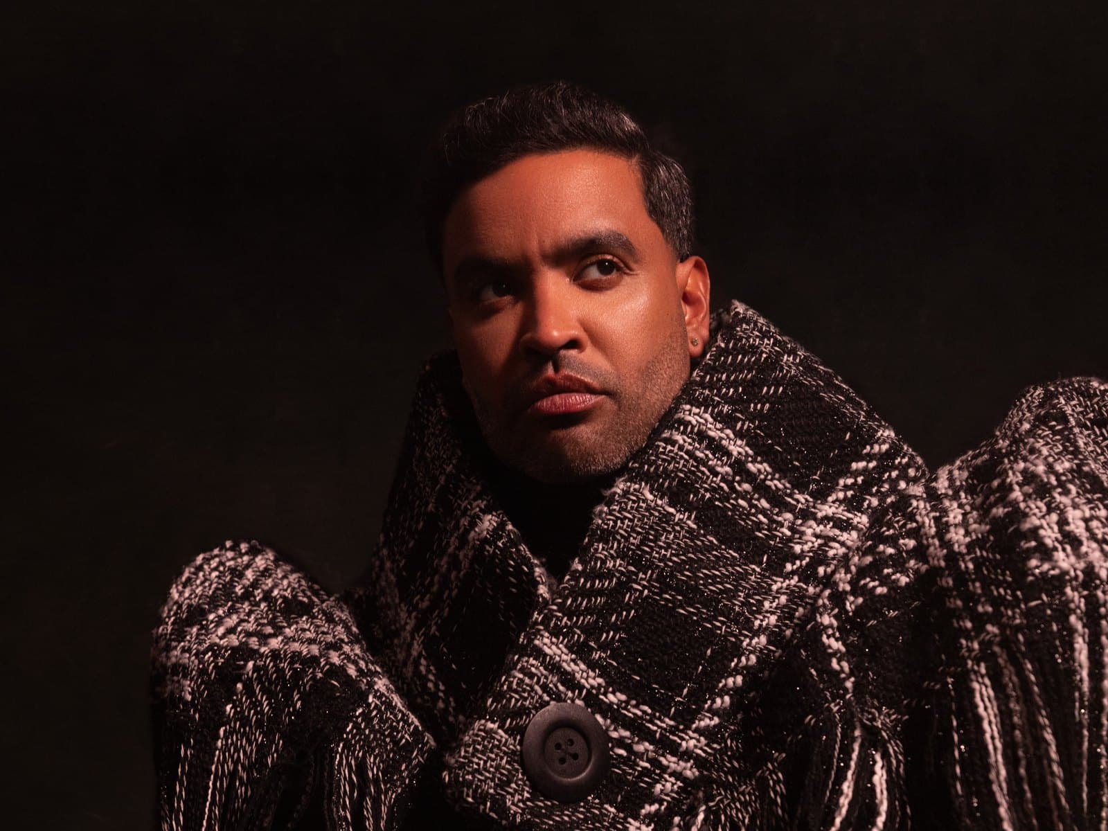 Seated portrait in a fringed black and white tweed shawl over a black knit, lit low against a dark ground