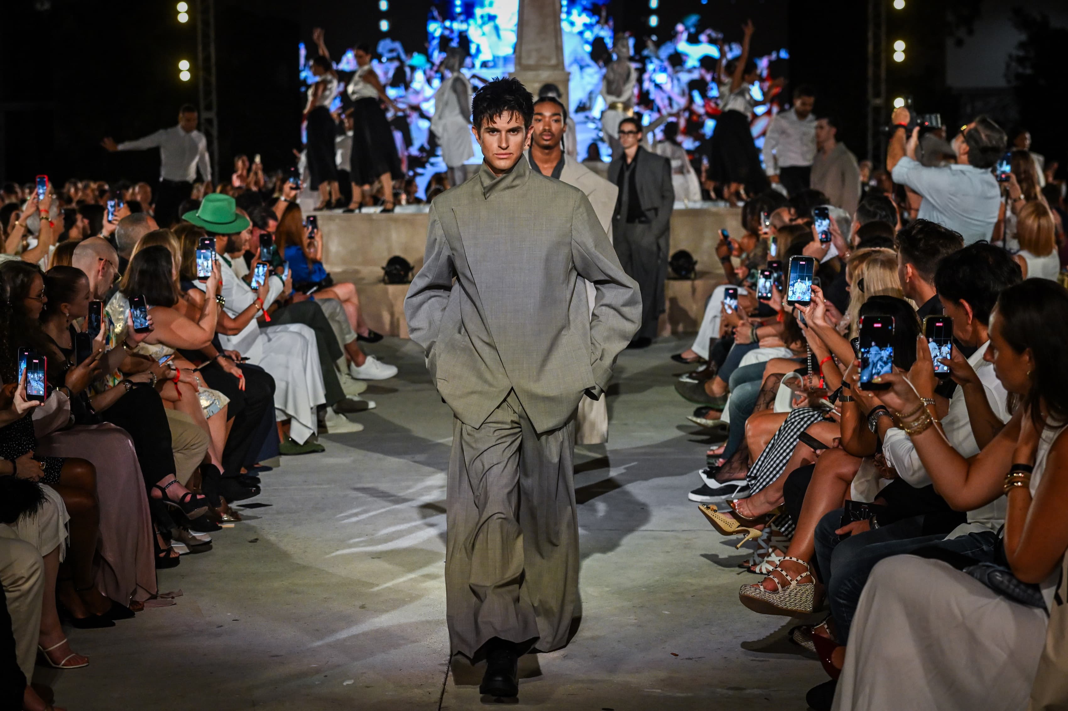 Runway photograph — a model in a grey high-collar suit with wide trousers walking the runway at night between rows of seated guests
