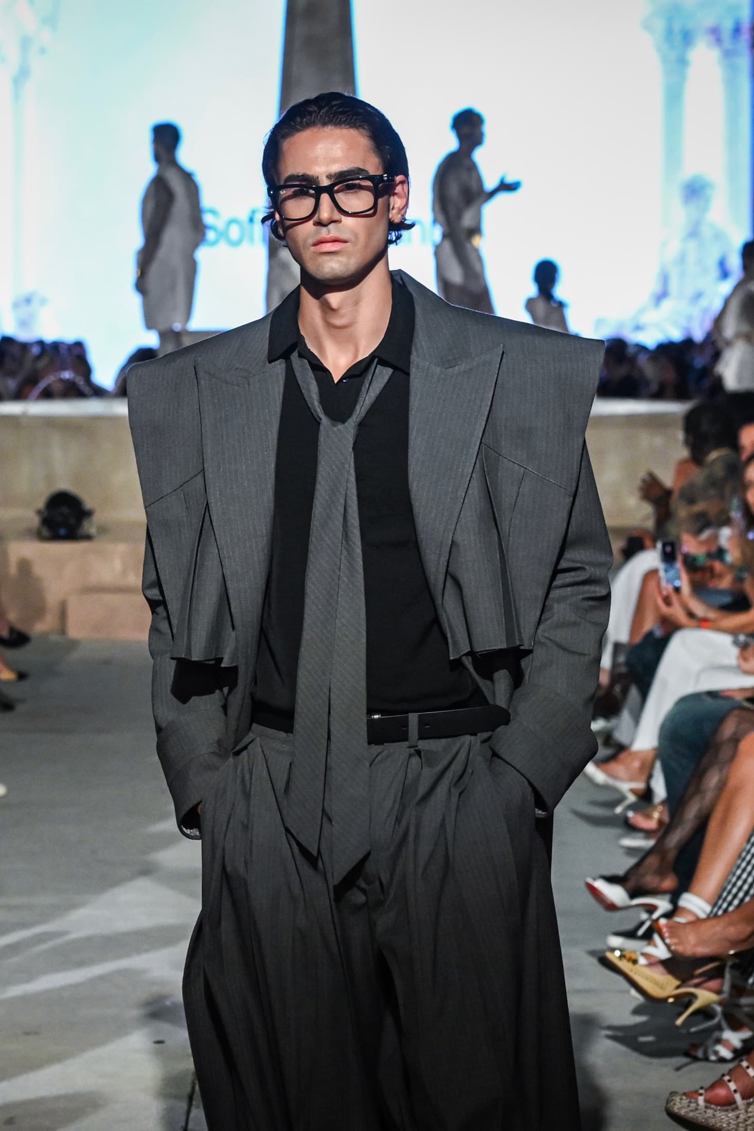 Model in a charcoal sculpted-shoulder suit with a grey tie and heavy-framed glasses — Miami Design District